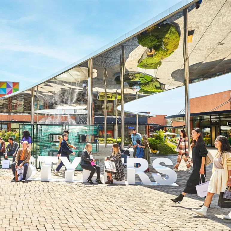 Persone che fanno shopping e si rilassano all’aperto a Vicolungo The Style Outlets, sotto una copertura riflettente e moderna